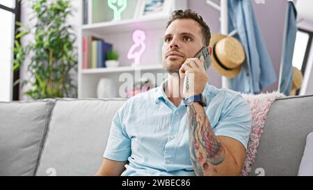 Un bell'uomo ispanico con barba e tatuaggi che parla al telefono in un soggiorno moderno Foto Stock