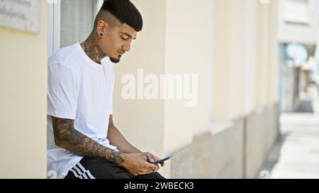 Un uomo latino alla moda che si avvale di uno smartphone e che digita testi con espressioni serene nello sfondo urbano e soleggiato Foto Stock