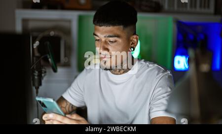 Bellissimo giovane latino, musicista tatuato, che esprime emotivamente la sua canzone sullo smartphone in mezzo alla notte buia nel suo studio musicale Foto Stock
