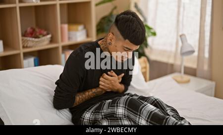 Bel giovane latino, tatuato e sofferente, seduto sul suo letto che stringe il petto in agonia per attacco di cuore a casa Foto Stock