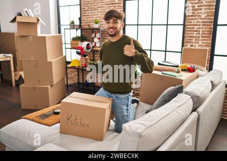 Uomo arabo con la barba che si sposta in una nuova casa chiudendo la scatola di cartone sorridente felice e positivo, pollice su facendo eccellente e segno di approvazione Foto Stock