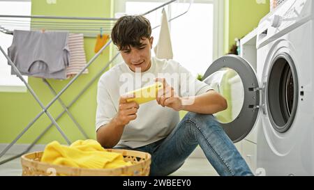 Colto nel bel mezzo delle pulizie, il giovane adolescente ispanico si impegna a giocare seriamente, aspettando vicino alla lavatrice della lavanderia Foto Stock
