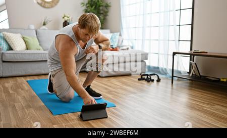 Un giovane biondo con la barba che usa un tablet mentre si esercita su un tappetino yoga in un soggiorno moderno. Foto Stock