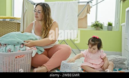 Madre e figlia condividono un momento serio mentre tengono insieme vestiti e un cestino di vimini nell'accogliente lavanderia della famiglia, un angolo della loro vigilia Foto Stock