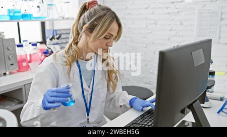 Una donna scienziata concentrata in un camice da laboratorio usa un computer mentre tiene un campione di liquido blu in un laboratorio ben attrezzato. Foto Stock