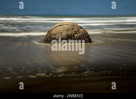 Bella scena artistica di un singolo masso rotondo, metà incastonato nella spiaggia, con dolci increspature d'acqua in primo piano e un mare a lunga esposizione sullo sfondo Foto Stock