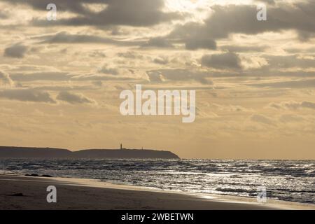 Questa fotografia presenta una tranquilla scena balneare mentre la giornata si conclude. La silhouette di un faro distante si erge come una sentinella all'orizzonte, affacciandosi sulle acque scintillanti baciate dal sole che tramonta. Le nuvole, sparse nel cielo, catturano la luce che si affievolisce, aggiungendo profondità e drammatismo alla scena. La spiaggia in primo piano offre un senso di calma solitudine, invitando lo spettatore a immaginare il suono delle onde e la sensazione di una leggera brezza. Questa immagine cattura l'essenza della bellezza costiera durante la magica ora del crepuscolo. Serenity costiero: Faro al tramonto. Fot. Di alta qualità Foto Stock