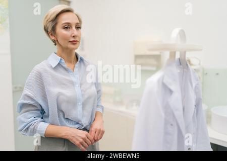 Un medico di mezza età pensivo entra in ufficio, per togliere l'abito dall'appendiabiti e metterlo. Professionista che si prepara a lavorare Foto Stock