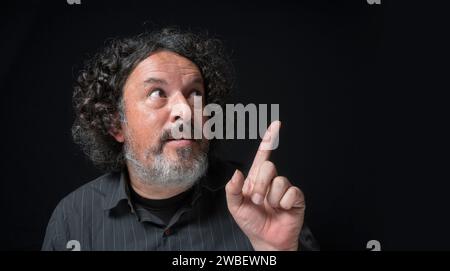 Ritratto di uomo latino con barba bianca e capelli ricci neri con divertente espressione, puntando verso l'alto con la mano, indossando una camicia nera contro lo schienale nero Foto Stock
