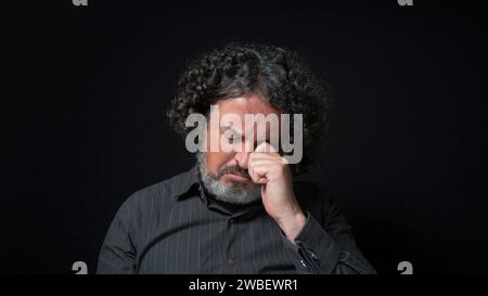 Ritratto di uomo latino con barba bianca e capelli ricci neri con triste espressione, occhi chiusi e testa giù, indossando una camicia nera contro lo sfondo nero Foto Stock