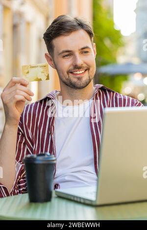 Uomo che usa carta di credito, computer portatile con carta di credito durante il trasferimento di denaro, acquista acquisti online, ordina la consegna di cibo, prenota la camera d'albergo. Un giovane turista seduto a un tavolo nella caffetteria della città. Verticale Foto Stock