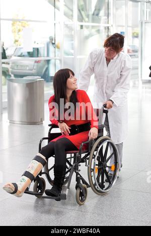 Young woman with plaster leg, wheelchair and carer Foto Stock