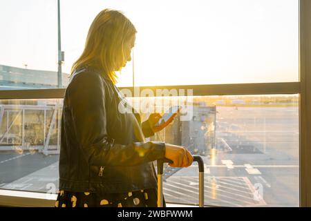 Pronta per l'imbarco, una giovane donna con una valigia controlla il suo smartphone. Foto Stock