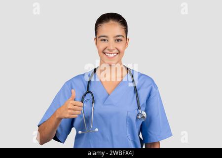 Medico millenario europeo fiducioso in scrub blu che dà un pollice in su Foto Stock