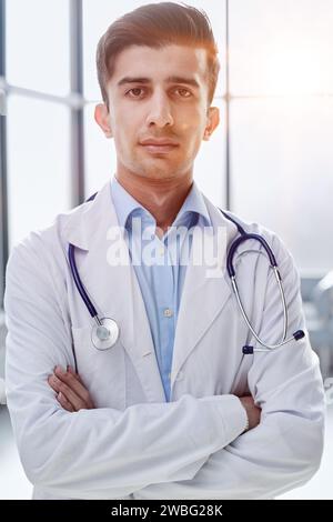 giovane medico sicuro di sé in uniforme bianca sorridente e guardando mentre si trova in piedi con le braccia incrociate Foto Stock
