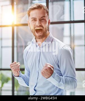Ritratto di un giovane uomo d'affari in una camicia blu che scuote i pugni celebrando il suo successo Foto Stock