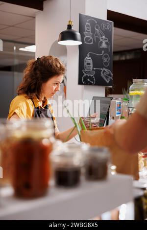 Magazzino presso il bancone cassa del supermercato zero rifiuti che utilizza uno scanner di codici a barre per elaborare le transazioni con il cliente. Venditore in un negozio di quartiere sostenibile che esegue la scansione di prodotti alimentari dei clienti Foto Stock