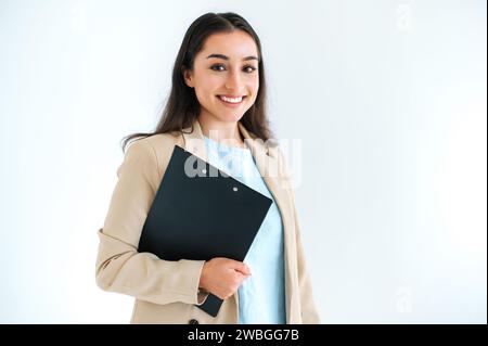 Donna d'affari indiana o araba di successo, consulente finanziario, manager, si trova su uno sfondo bianco isolato, tiene una cartella con i documenti, guarda la fotocamera e sorride Foto Stock