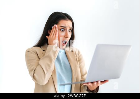 Scioccata, confusa donna d'affari indiana o araba, dipendente in ufficio, con un portatile aperto, frustrato lo guarda con la bocca aperta, in piedi su uno sfondo bianco isolato, afferrando la testa con la mano Foto Stock