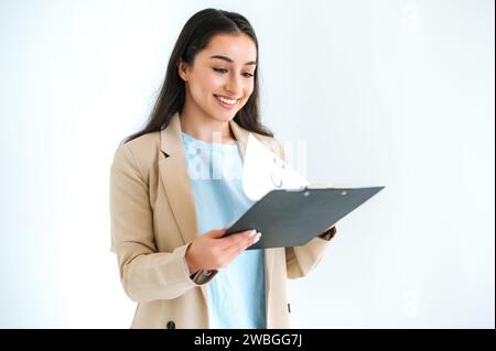Sicura di sé, donna d'affari indiana o araba, consulente finanziario, commerciante, si trova su sfondo bianco isolato, tiene una cartella con documenti e grafici, guardandola, sorride Foto Stock
