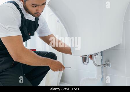 In un bagno un tecnico ripara un tubo dell'acqua sotto il lavandino. L'esperto idraulico si concentra su Foto Stock