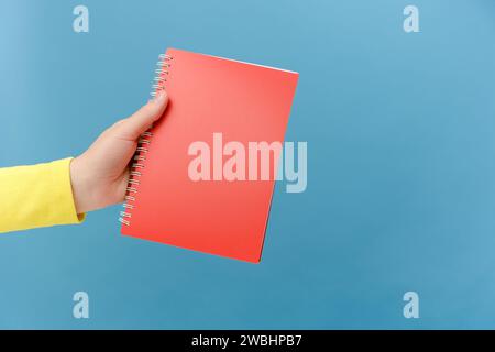 Primo piano della mano di una donna che tiene un quaderno bianco rosso, spazio vuoto per il testo commerciale, area pubblicitaria su carta, posa isolata su blu semplice b Foto Stock