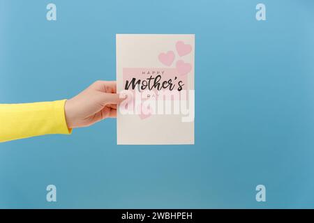 Primo piano della carta di mano femminile per la Festa della Mamma con piccoli cuori rosa, in posa isolata su una parete di sfondo di colore blu in studio con spa copiatrice Foto Stock