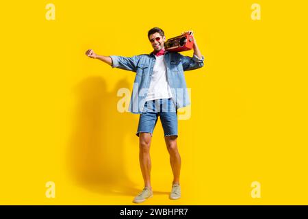 Foto integrale di pantaloncini camicia in denim vestito da uomo soddisfatto con occhiali da ballo con stereo isolato su sfondo giallo brillante Foto Stock