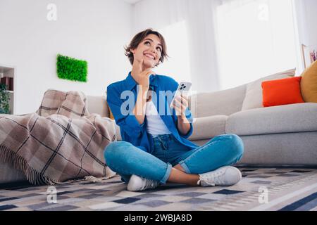Foto di una ragazza sognante e carina seduta sul pavimento in un appartamento interno che sceglie di prenotare un hotel per il fine settimana Foto Stock