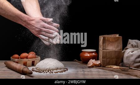 Baker sta cucinando del pane. Batti le mani con la farina sull'impasto. Farina nell'aria. Cucina fatta in casa. Sul tavolo della cucina c'è un cesto di uova, un ba Foto Stock