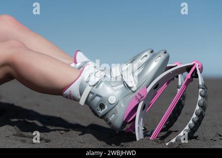 Closeup gambe femminili sottili in Kangoo salta stivali sulla spiaggia di sabbia nera mentre riposi durante l'allenamento aerobico e fitness. Vista laterale Foto Stock