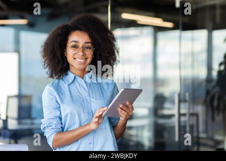Una giovane e bella donna d'affari con i capelli ricci che lavora in ufficio, tenendo in mano un tablet. Foto Stock