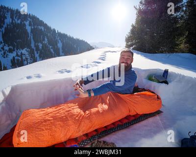 Viaggiatore mattutino in montagna in inverno. Un viaggiatore con la barba ha passato la notte in un sacco a pelo in un rifugio sulla neve Foto Stock