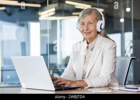 Ritratto di una donna d'affari anziana e matura che si trova sul posto di lavoro all'interno dell'ufficio, donna sorridente e che guarda la fotocamera che lavora con un notebook, ascolta podcast e audiolibri, musica in cuffia. Foto Stock
