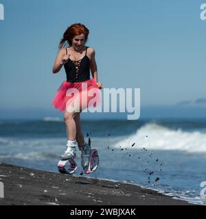 KAMCHATKA, RUSSIA - 15 GIUGNO 2022: Sportiva nello sport Kangoo salta stivali, costume da bagno monopezzo nero e gonna corta rosa che salta e corre sulla sabbia Foto Stock