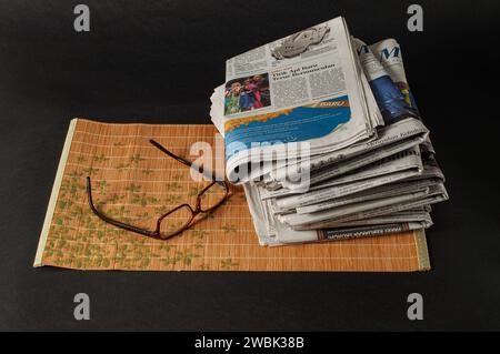 pile di giornali da leggere per notizie e informazioni finanziarie e occhiali per una maggiore chiarezza, fotografati su uno sfondo scuro Foto Stock