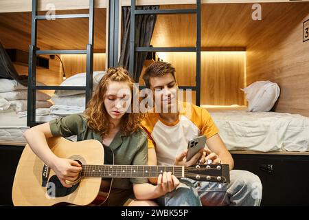 giovane donna che suona la chitarra acustica al fidanzato con lo smartphone in una stanza accogliente dell'ostello degli studenti Foto Stock