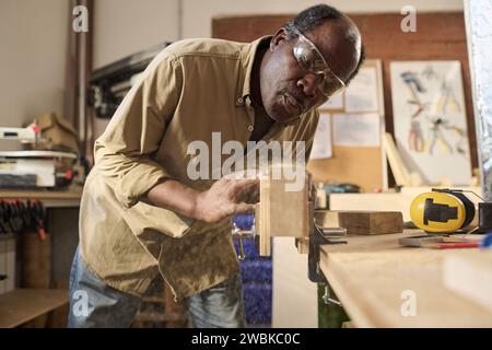 Ritratto di un falegname nero che soffia la segatura su un asse di legno mentre costruisce mobili in officina Foto Stock