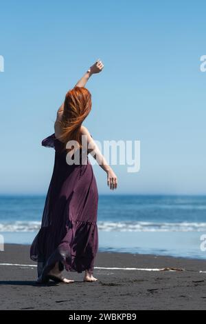 Donna rossa con abito lungo e scuro in piedi su una spiaggia sabbiosa con la mano sollevata, sullo sfondo del mare blu cristallino nelle soleggiate giornate estive con cielo limpido Foto Stock