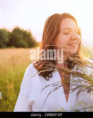 Giovane donna incinta sinceramente sorridente vestita con abiti estivi leggeri che passeggia accanto all'alto prato verde con bouquet di fiori selvatici nei soli serali Foto Stock