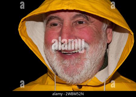 Il vecchio pescatore con barba barba, giacca impermeabile gialla e cappuccio ride, mentre le rughe intorno agli occhi rivelano una storia di vita piena di esperienze. Foto Stock