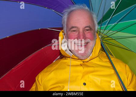 Di buon umore nonostante il maltempo, un anziano sorridente con una giacca impermeabile gialla e un ombrello colorato irradia gioia. Foto Stock