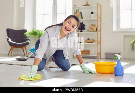 Giovane donna felice che pulisce la polvere con spray e straccio mentre pulisce il pavimento a casa. Foto Stock