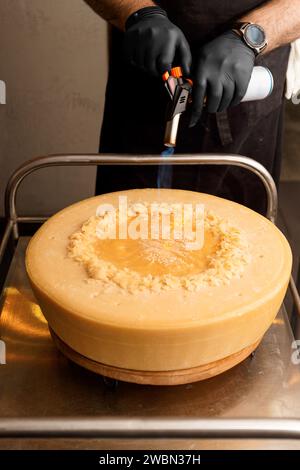 Lo chef scioglie il formaggio utilizzando il calore di una torcia a gas, preparando la ruota di parmigiano per la presentazione e la porzione di pasta Foto Stock