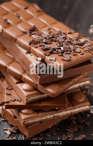 Una pila di barrette di cioccolato intere e rotte e gocce di cioccolato sparse su uno sfondo di legno. Diversi tipi di cioccolato sono impilati in una pila Foto Stock