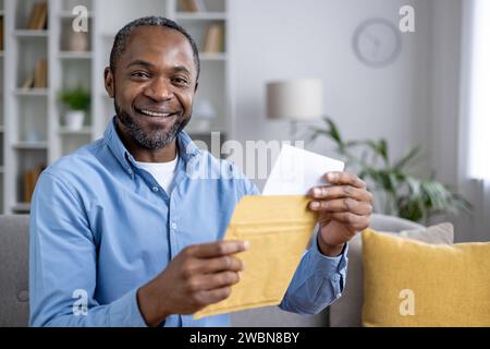 Ritratto ravvicinato di un uomo afro-americano sorridente seduto a casa e con una busta aperta con una lettera in mano, che guarda e sorride la fotocamera. Foto Stock