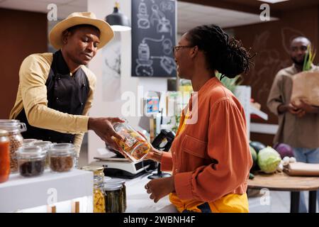 L'uomo e la donna afroamericani discutono di alimenti freschi e nutrienti. Al banco cassa, una cliente donna consegna il barattolo di pasta al fornitore maschio della cassiera per la scansione. Foto Stock