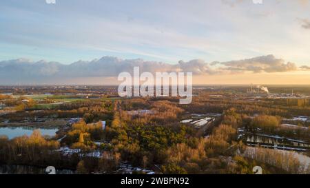 Questa immagine offre una prospettiva aerea di un paesaggio lussureggiante e verde che si trasforma in uno skyline industriale. Il primo piano è un mosaico di foresta e acqua, una testimonianza della resilienza dell'ambiente naturale. Mentre l'occhio si sposta verso l'orizzonte, la scena si sposta verso la feroce funzionalità delle strutture industriali, con pennacchi di vapore o fumo che si innalzano contro il cielo. Il contrasto tra le due zone è colmato dalla luce soffusa di quello che sembra essere un tramonto o un sole nascente, accennando al continuo ciclo del giorno e della notte su sforzi umani e ambientazioni naturali allo stesso modo.Hori industriale Foto Stock