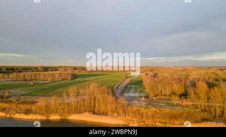 Questa immagine aerea cattura un paesaggio rurale immerso nel caldo bagliore di un sole che tramonta. Una strada tortuosa attraversa la scena, che conduce all'orizzonte, fiancheggiata da campi in vari stati di utilizzo, alcuni portano i segni di aratura recente, mentre altri sono rigogliosi di colture. Un corso d'acqua si snoda lungo il bordo dei campi, riflettendo la luce del sole e aggiungendo un elemento dinamico alla campagna altrimenti ancora incontaminata. Macchie di alberi punteggiano il paesaggio, fornendo un habitat per la fauna selvatica e una pausa nella distesa di campi aperti. Lo skyline distante è segnato da una soffice foschia, probabilmente il risultato dell'oro Foto Stock