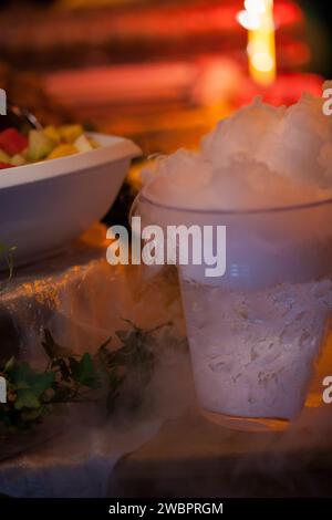 Questa accattivante immagine cattura un punch fresco avvolto da una mistica nebbia, probabilmente causata dal ghiaccio secco, durante un banchetto festivo. La nebbia aggiunge un elemento di intrigo e teatralità alla scena, mentre si riversa sul tavolo, creando un effetto etereo. Sullo sfondo, l'illuminazione calda e soffusa e il contorno sfocato di una ciotola di frutta contribuiscono all'atmosfera conviviale del raduno. La fotografia suggerisce un evento pieno di delizie sensoriali, dove la presentazione di cibo e bevande fa parte dell'esperienza. Mist mistico: Delizie fresche in un banchetto festivo. Pho di alta qualità Foto Stock